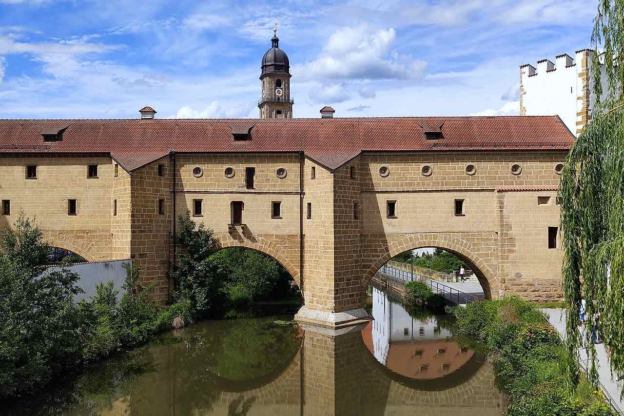 The Stadtbrille, a sandstone city-wall building with two arches over the river Vils that mirror in the water, with the tower of the St. Martin basilica behind it