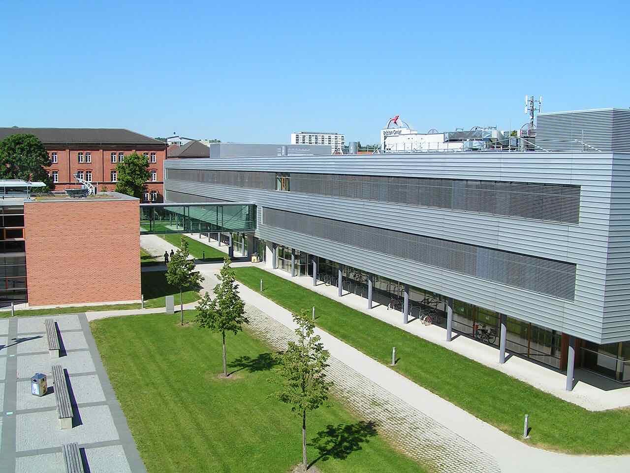 A long modern grey-clad building with a glass bridge, lawns and a red-brick wing on the Hochschule Ansbach campus under a blue sky