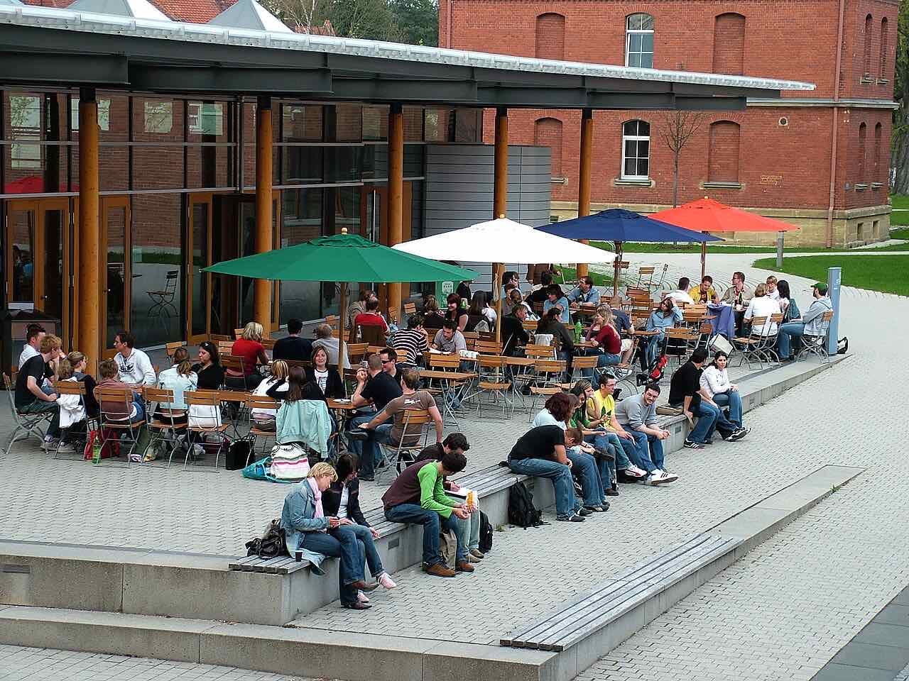 Students sitting at tables under coloured parasols and on stone steps outside the canteen on the Hochschule Ansbach campus, with an old red-brick barracks building behind