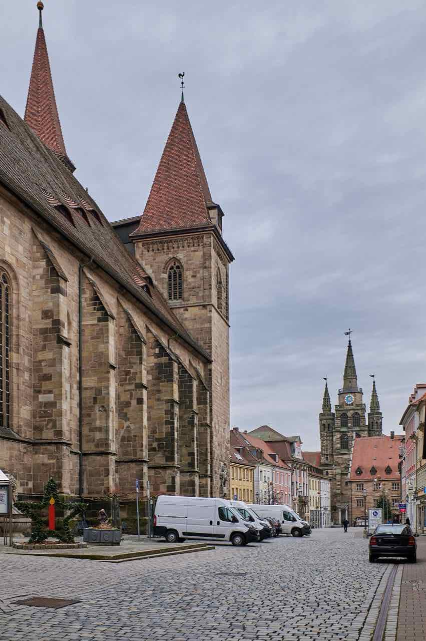 The sandstone side of St. Johannis church with a red pointed roof on a cobbled street, and the three spires of St. Gumbertus in the distance