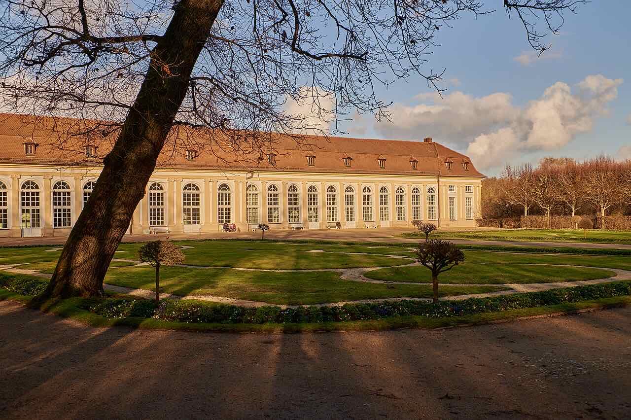 The long Orangerie in the Ansbach Hofgarten in late afternoon light, with arched windows, a lawn with clipped trees in front and a bare tree in the foreground