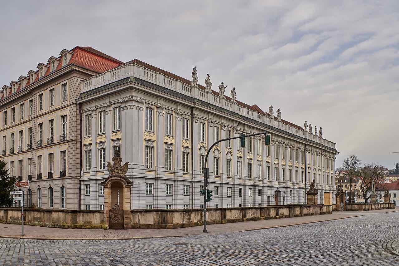 The long cream and grey Baroque front of the Ansbach Residenz with statues along the roof line, seen from a cobbled street corner under a cloudy sky