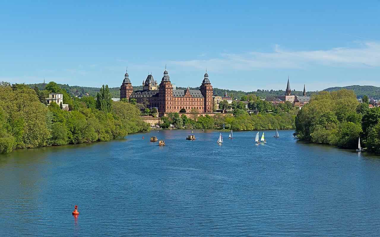 The red sandstone Renaissance palace with four towers on the far bank of the wide, blue River Main, with small sailing boats on the water and wooded hills behind