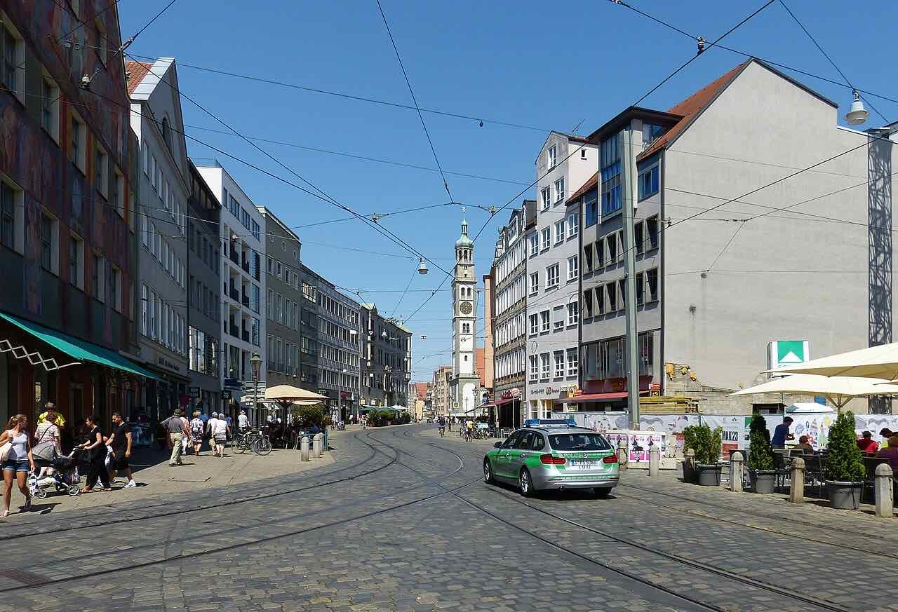 Maximilianstraße in Augsburg on a sunny day, with tram tracks, cafe terraces and the tall Perlach Tower at the end of the street
