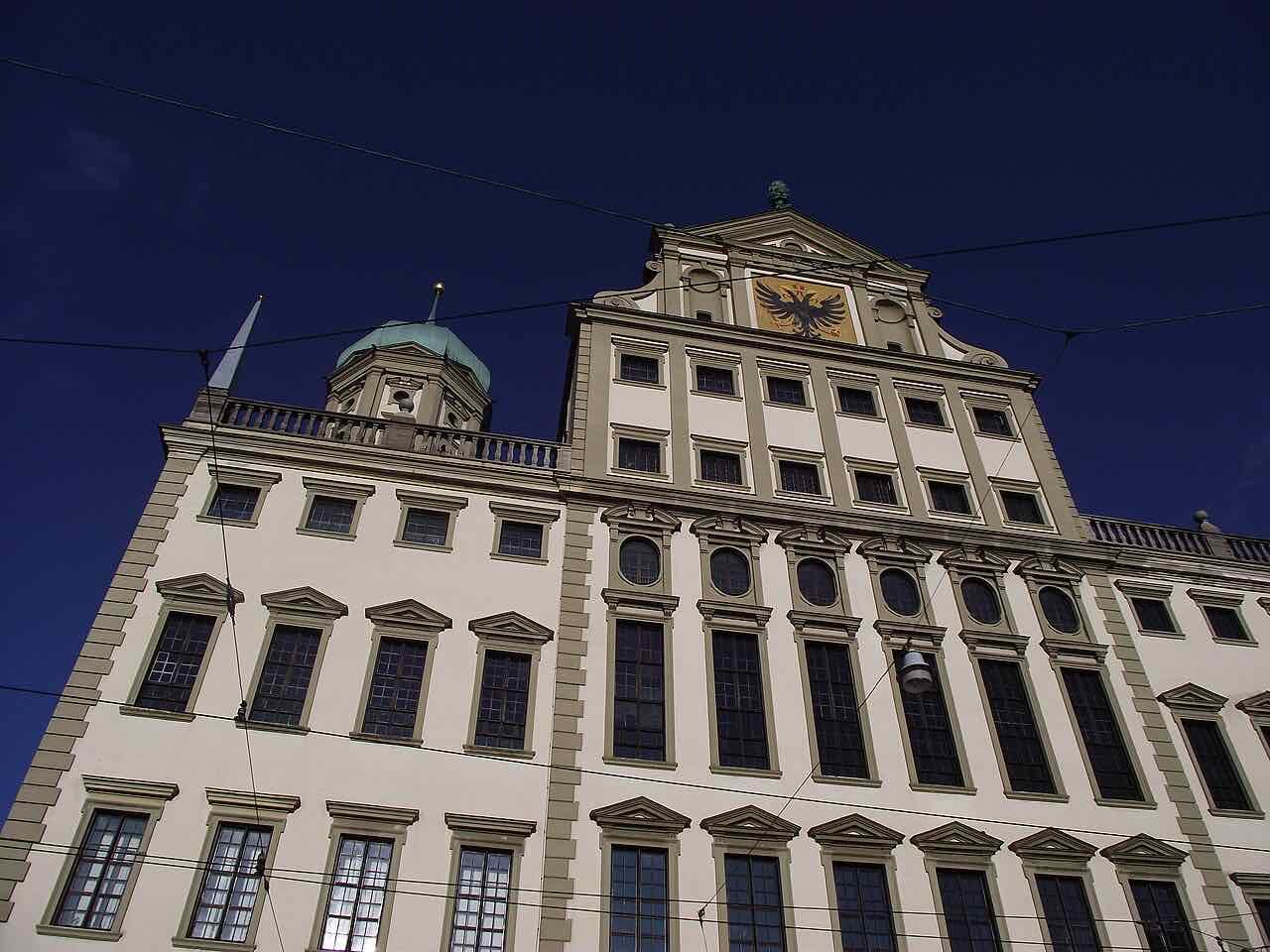 The Renaissance front of the Augsburg town hall seen from below against a deep blue sky, with a green copper dome and the golden eagle of the city