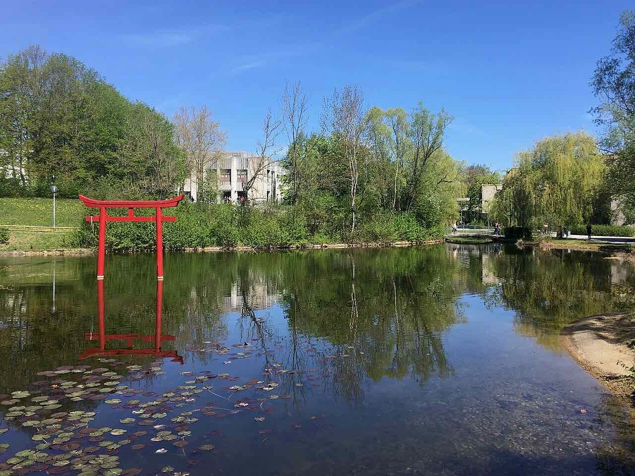 The pond on the University of Augsburg campus on a sunny day, with a red torii gate standing in the water and the concrete university buildings behind the trees