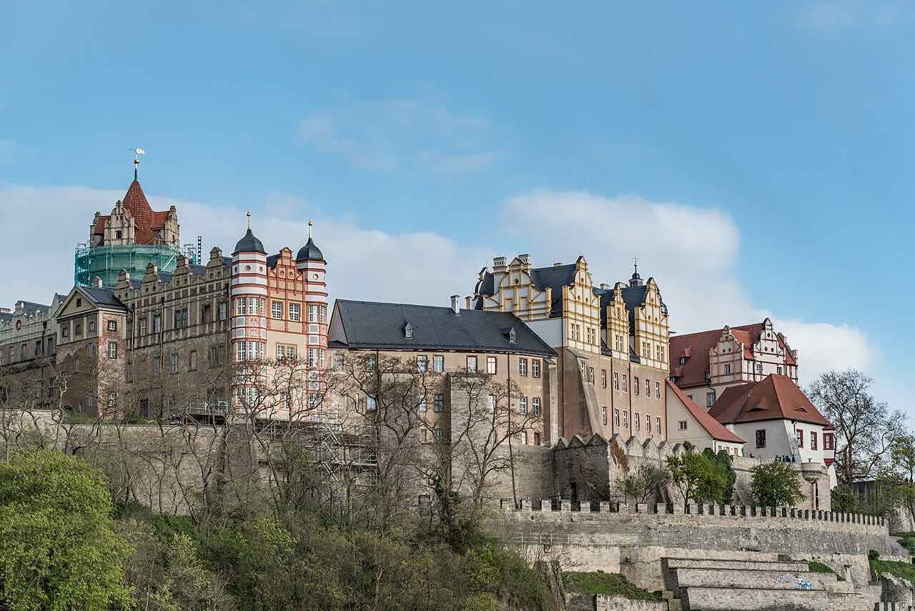 Bernburg castle above the Saale seen from the river side, with Renaissance gables, round corner towers and a stone retaining wall under a blue sky