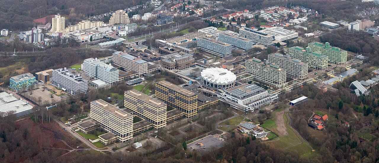 Aerial view of the Ruhr University Bochum campus with its concrete lecture buildings, the white Audimax dome in the middle and forest around the edges