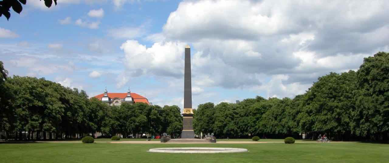 A wide lawn at the Löwenwall with a tall black obelisk and tall trees on both sides