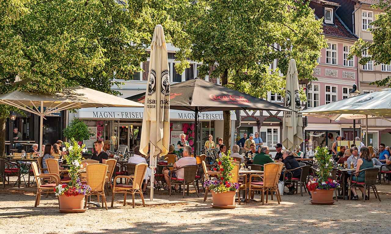 Café tables under trees on a gravel square in the Magniviertel, with pastel and half-timbered house fronts behind