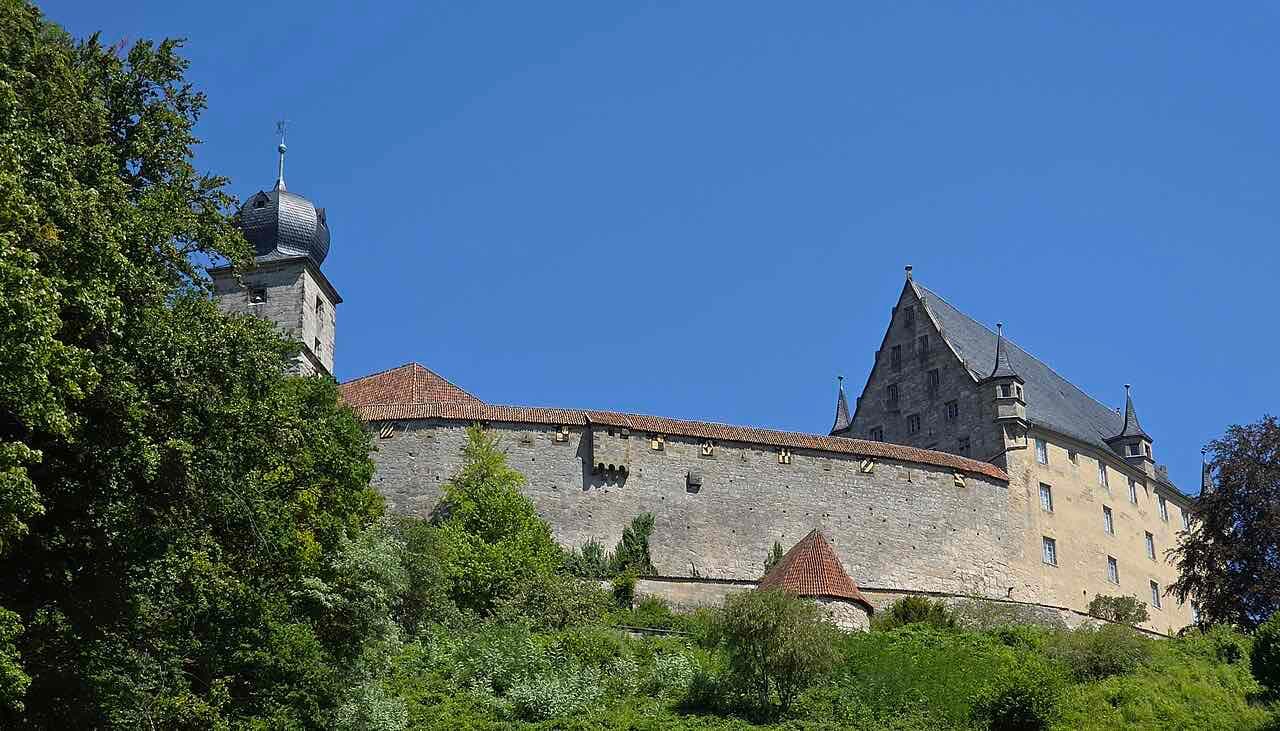 The grey stone wall, round tower and steep slate roofs of Veste Coburg seen from below through green trees under a clear blue sky