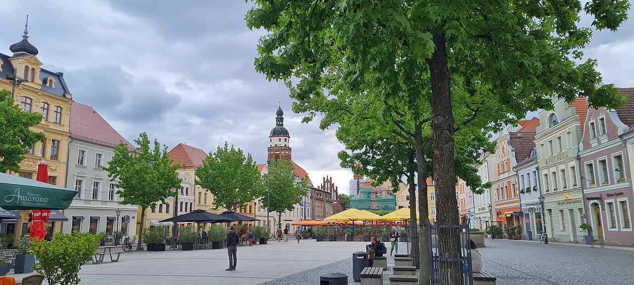 The Altmarkt in Cottbus on a cloudy day, with pastel-coloured town houses, café umbrellas and the brick tower of the Oberkirche behind the trees