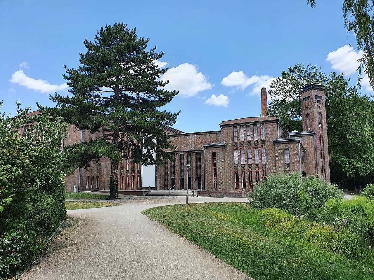 The red-brick Dieselkraftwerk in Cottbus with a tall chimney and tower, seen from a gravel path in a park with a large pine