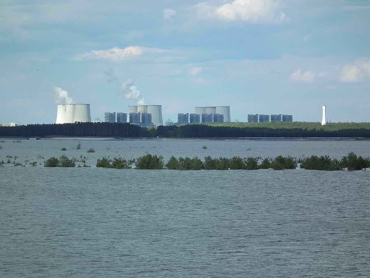 The wide water of the Cottbuser Ostsee with small shrubs standing in the shallows and the cooling towers of a power station on the far shore