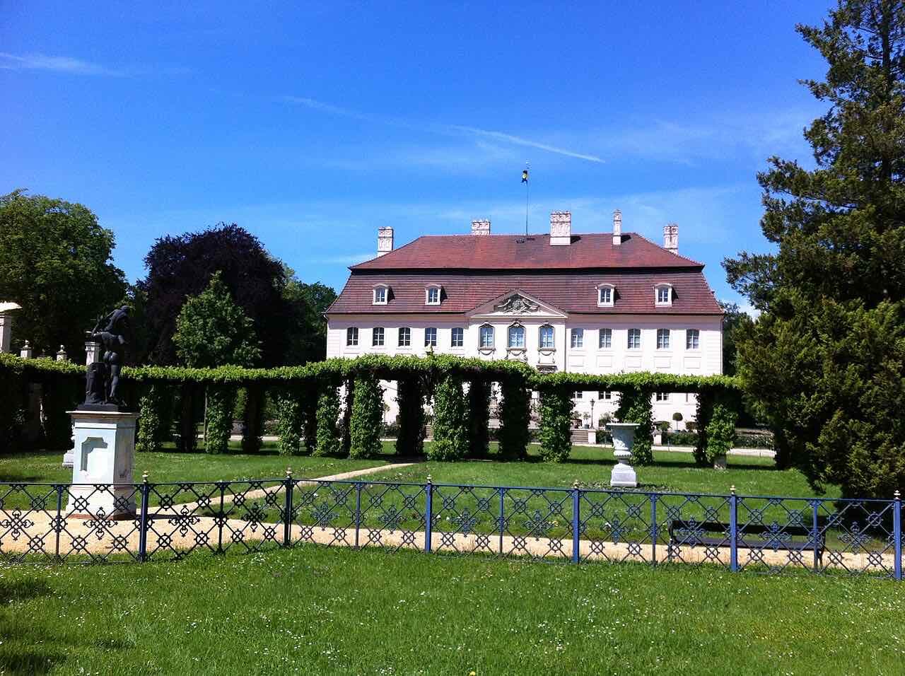 Schloss Branitz seen across a lawn and a green pergola, a cream-coloured palace with a red roof under a blue sky