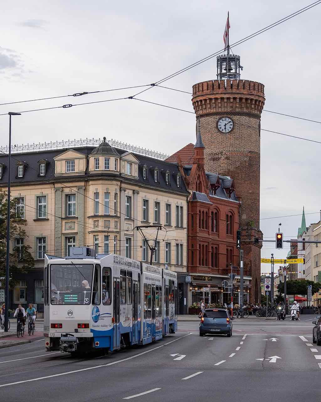 A blue and white tram passing the round brick Spremberger Turm with its clock, next to a red-brick building and a cream-coloured town house
