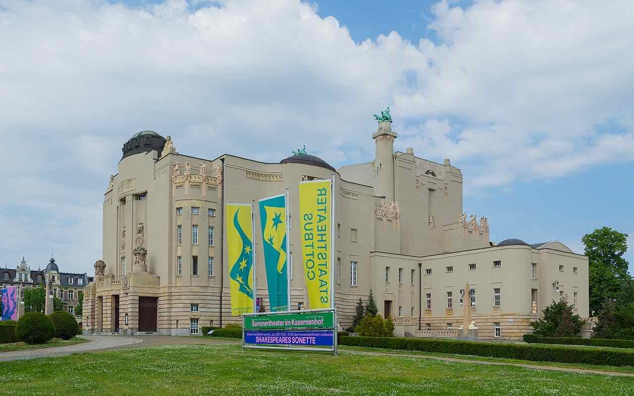 The Staatstheater Cottbus, a large pale stone theatre building with sculpted figures on its roofline and yellow and green banners in front