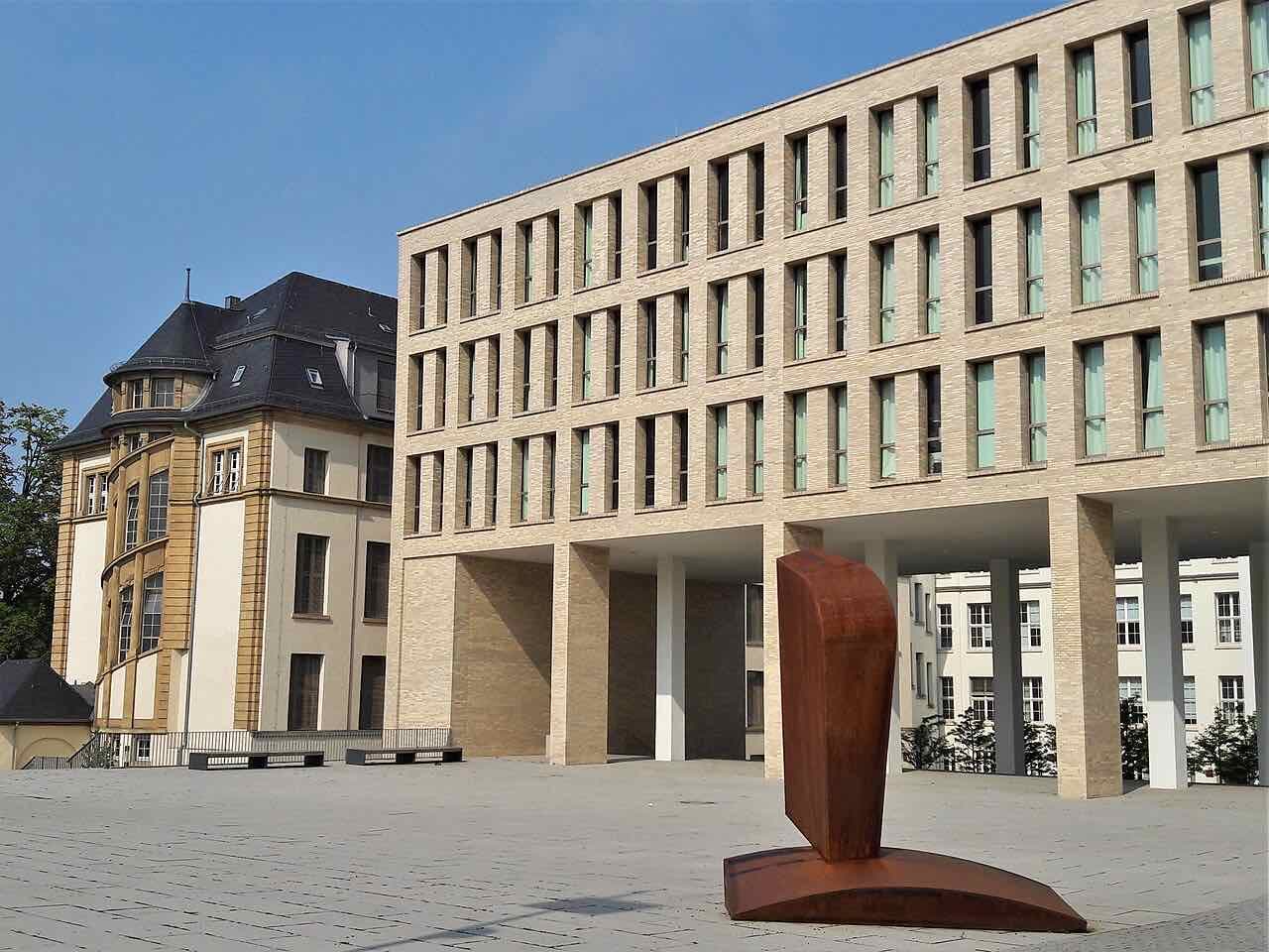 A modern light-brick university building on a paved square at TU Darmstadt, with an older yellow building at left and a rusty steel sculpture in front
