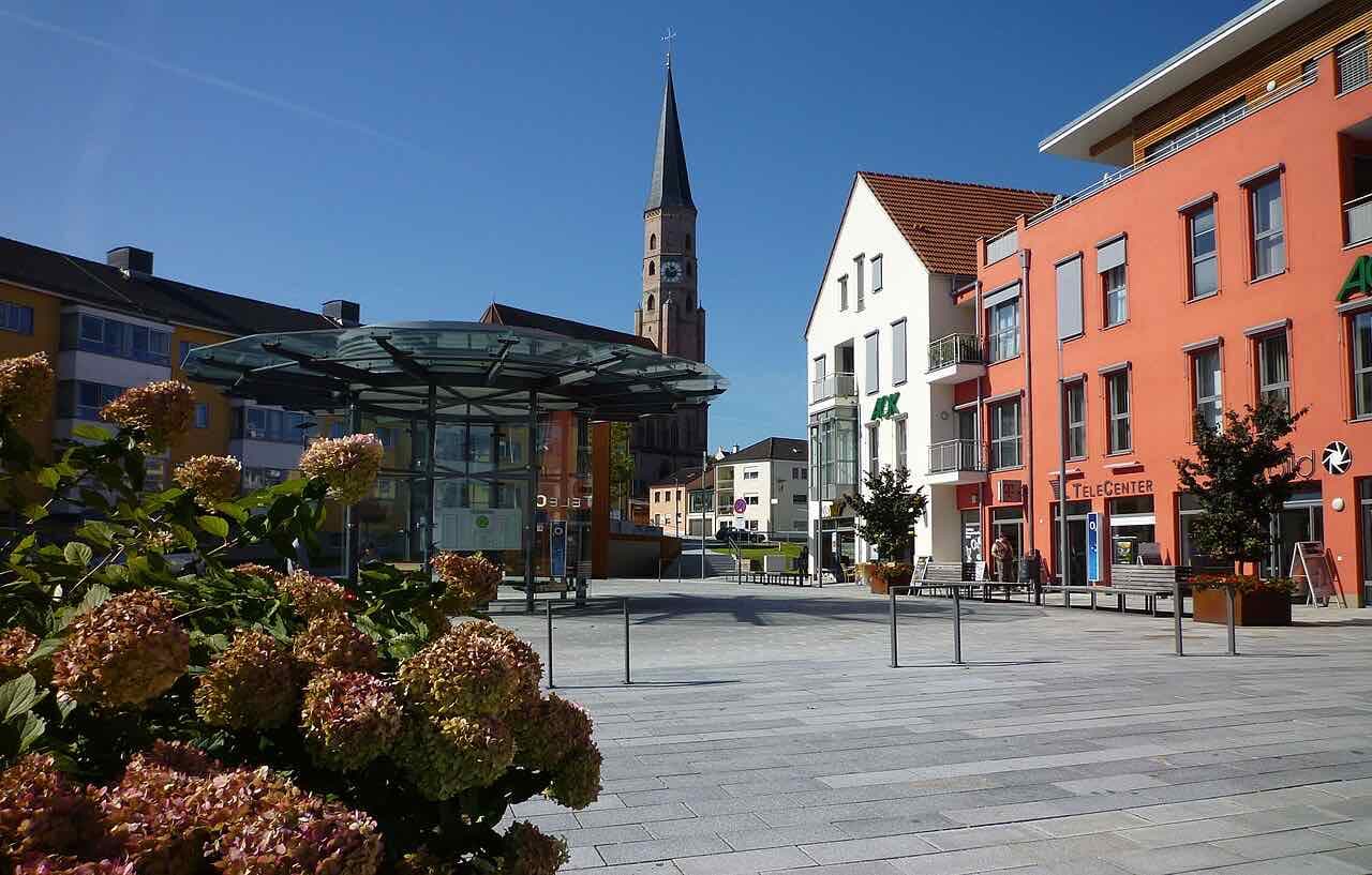 A wide paved square under a deep blue sky with a glass bus shelter, an orange-red and a white modern building on the right, hydrangeas in the foreground and the pointed brick tower of the St. Johannes church rising behind the shelter