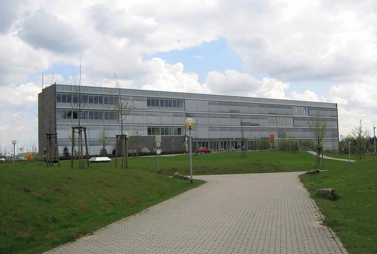 A long grey-clad building with a flat roof behind a lawn and a paved path under a cloudy sky