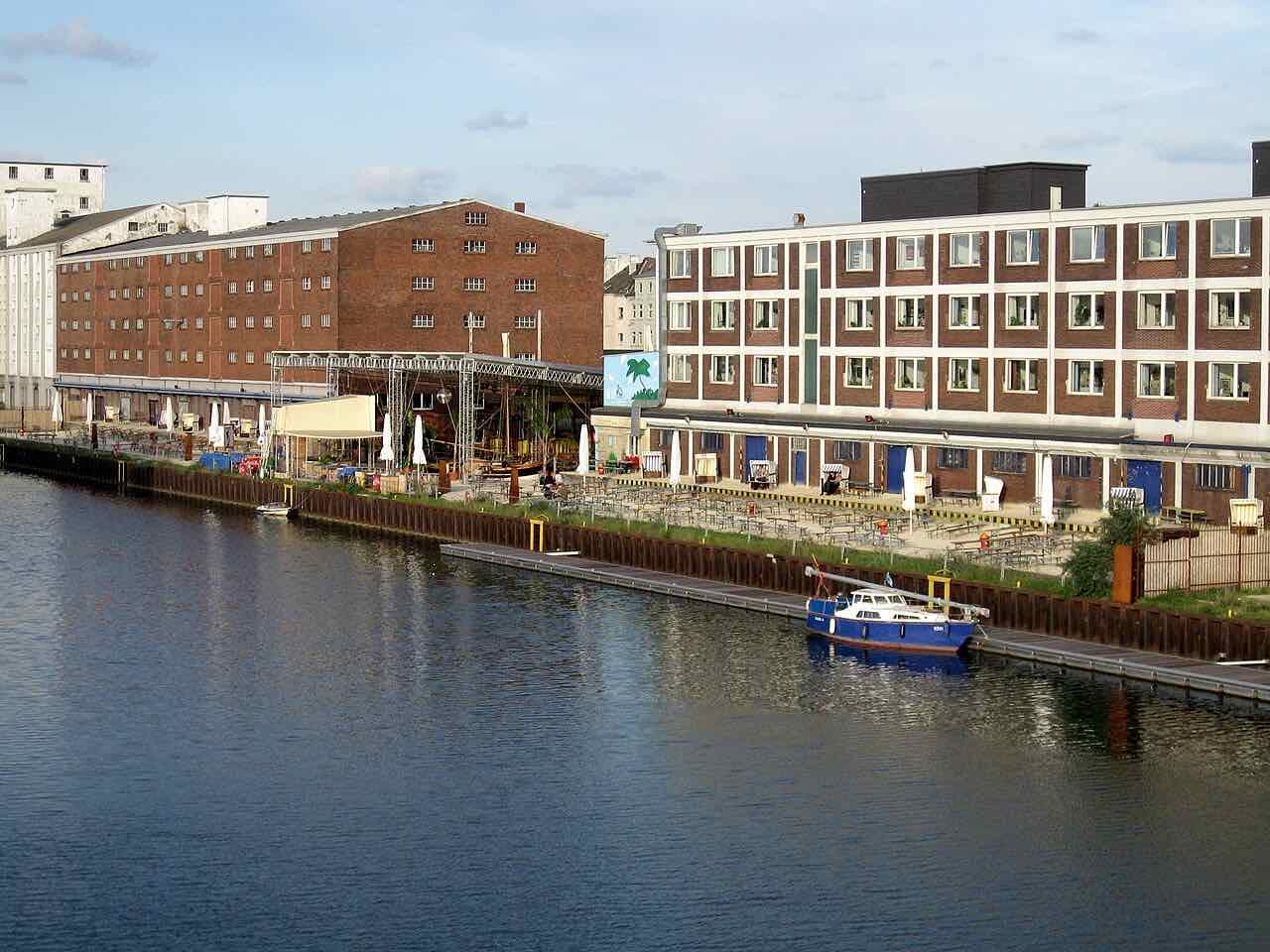 Brick warehouses and a four-storey block along the Dortmund harbour basin, with outdoor seating on the quay and a blue boat moored in front