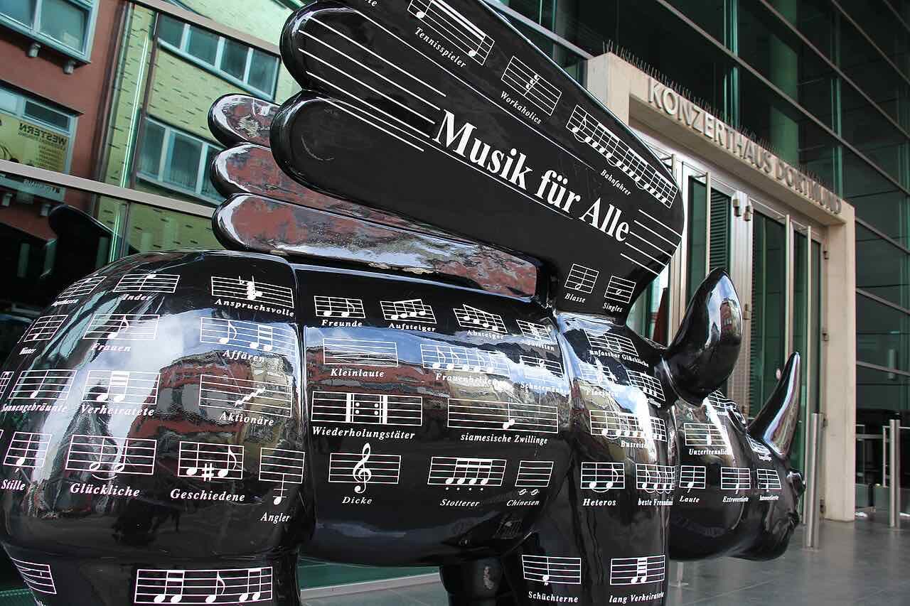 A black winged rhinoceros sculpture covered in music staves and the words Musik für Alle, standing in front of the Konzerthaus Dortmund entrance