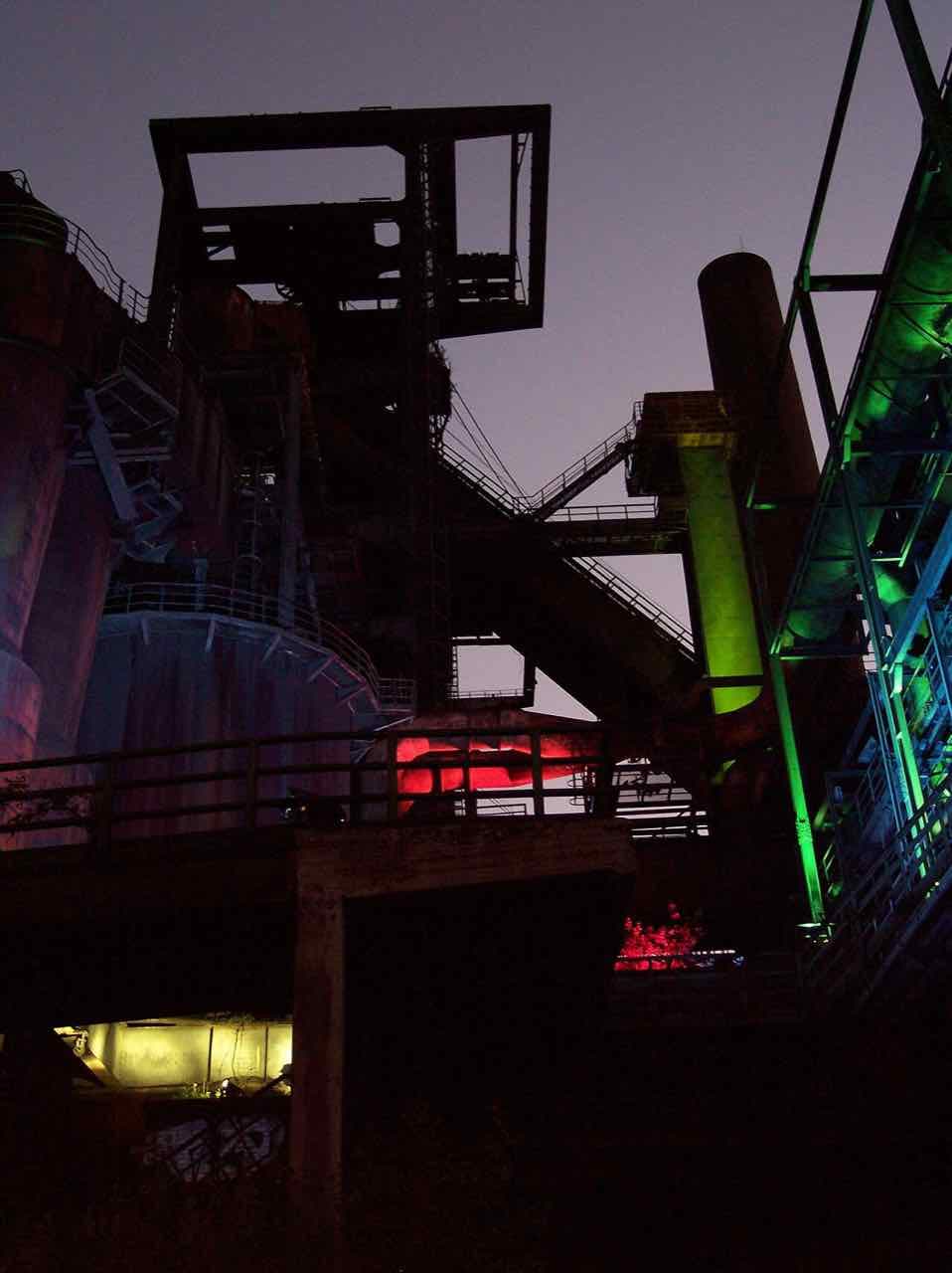 Blast furnace structures at night with stairways and pipes lit in red, green and blue
