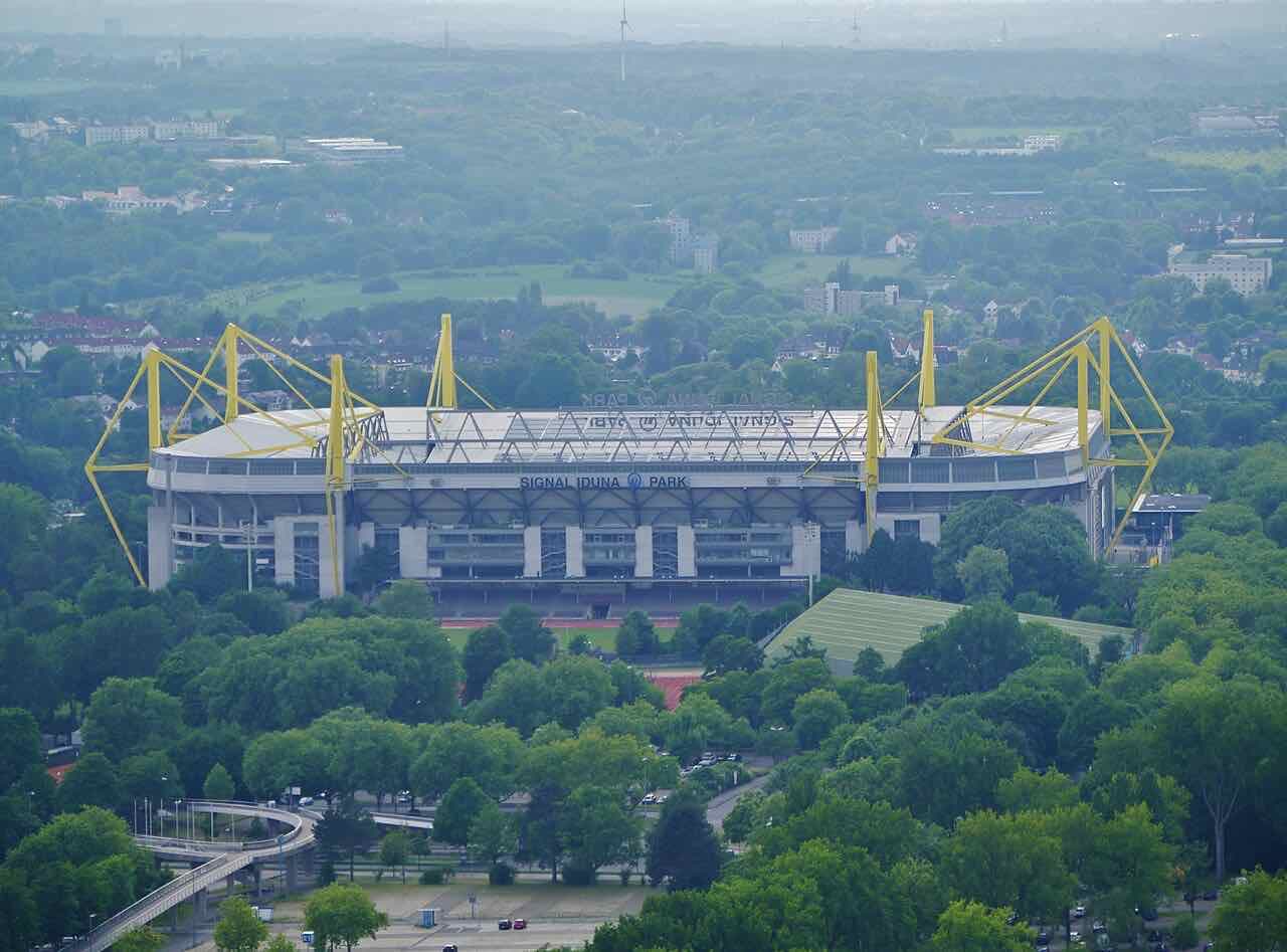 The yellow steel arches of the Signal Iduna Park stadium seen from above, surrounded by green treetops