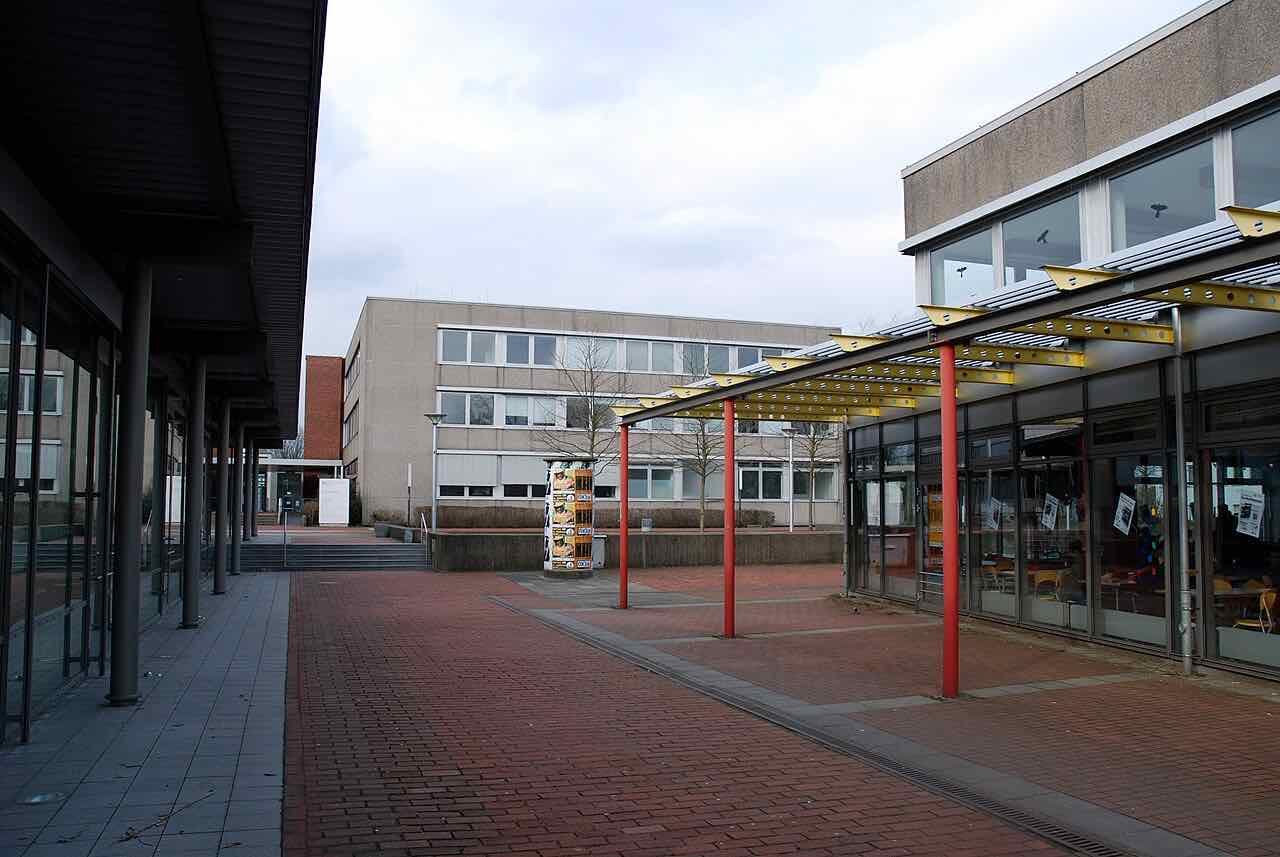 A brick-paved square between low concrete university buildings, with red steel posts carrying a yellow canopy in front of a cafeteria