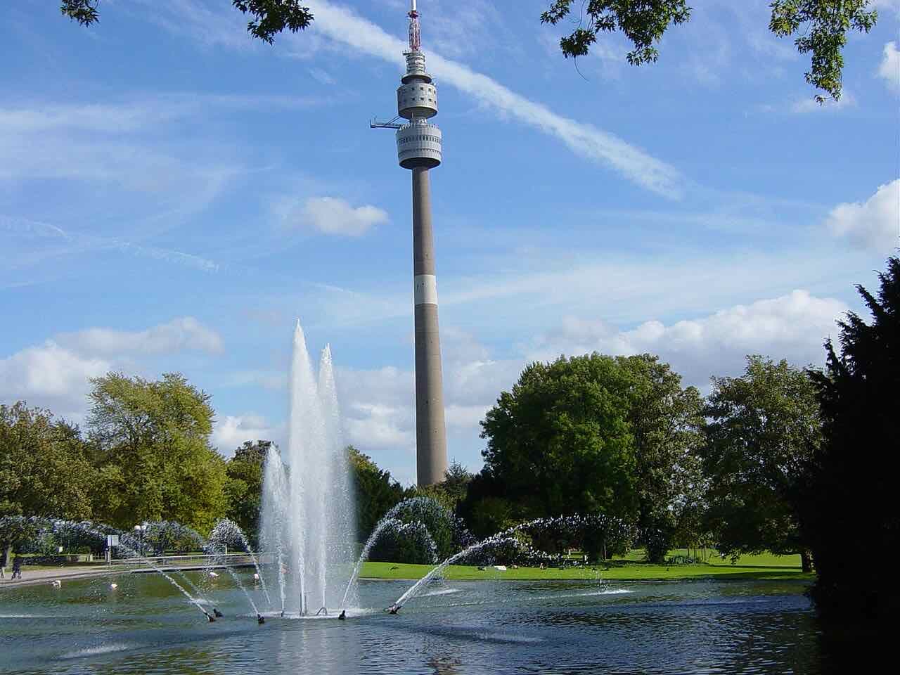 The Florianturm television tower rising above trees, with a fountain playing in a pond in the foreground