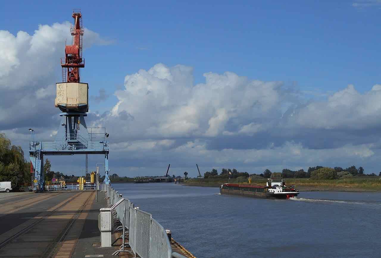 A red and blue harbour crane on a quay beside a wide grey river, a cargo barge moving along the water and the dark shapes of a flood barrier in the distance under a big cloudy sky
