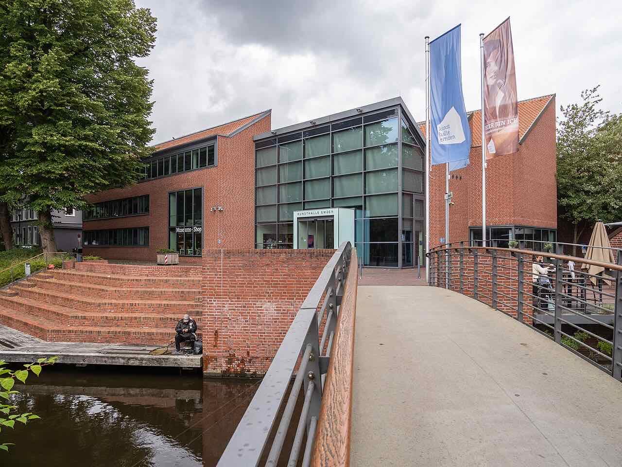 The red-brick and glass Kunsthalle Emden seen from a footbridge, with steps down to the town moat where a man is fishing