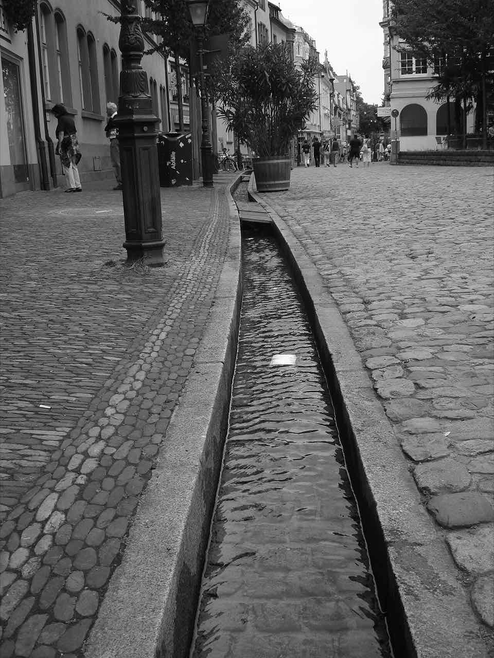 A narrow water channel, a Bächle, running along a cobbled street in Freiburg old town in black and white