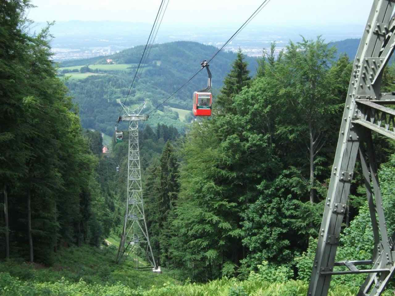 A red gondola of the Schauinslandbahn cable car above dark conifer forest, with the Rhine plain hazy in the distance
