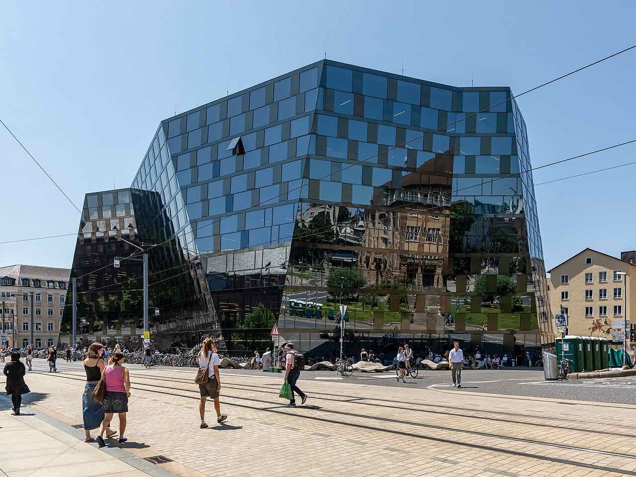 The mirrored glass University Library of Freiburg on a sunny day, with tram tracks and students crossing the square in front