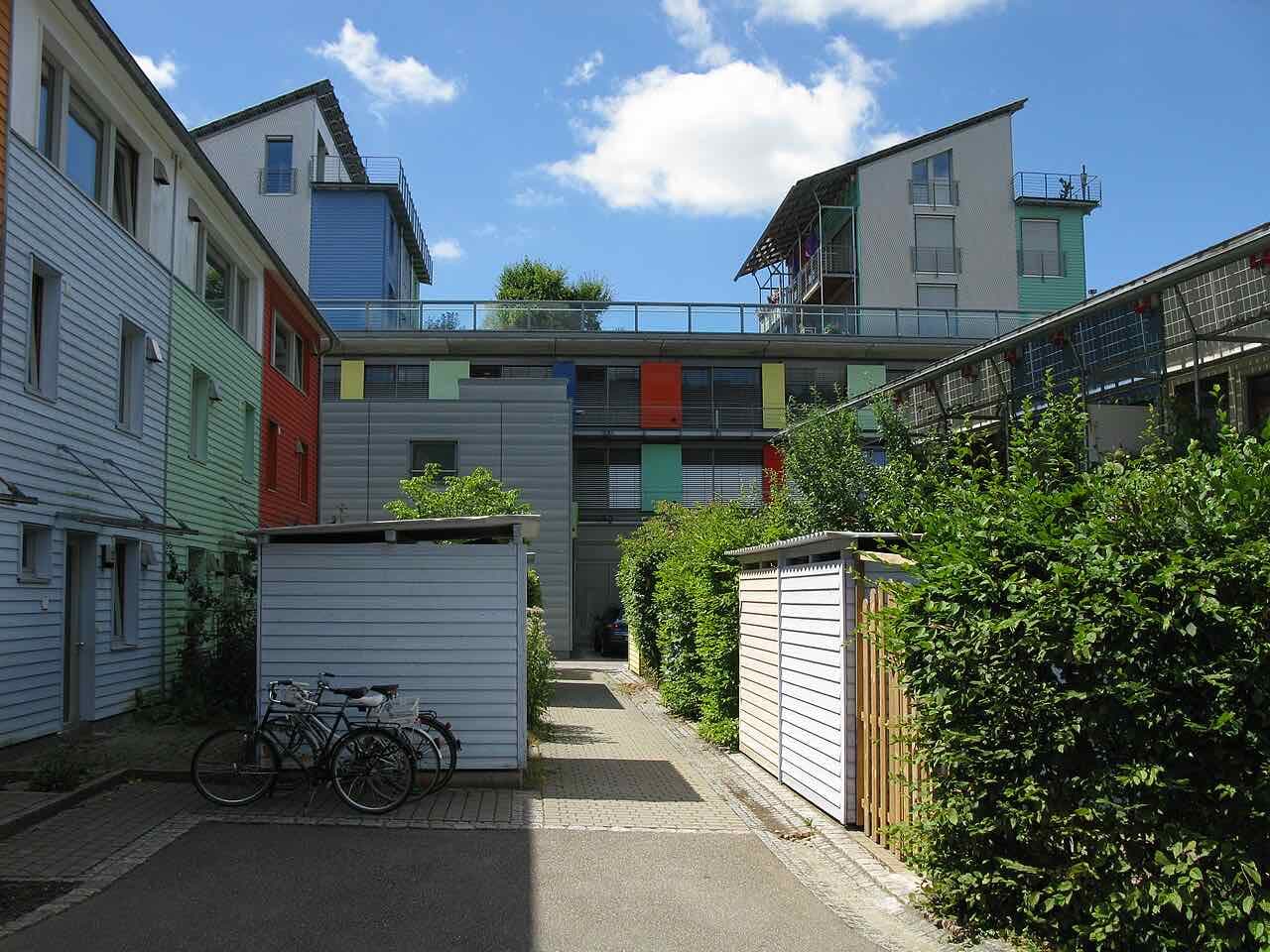 Colourful timber-clad houses and a solar-roofed building in the Vauban district, with bicycles parked beside a sheds and a green hedge