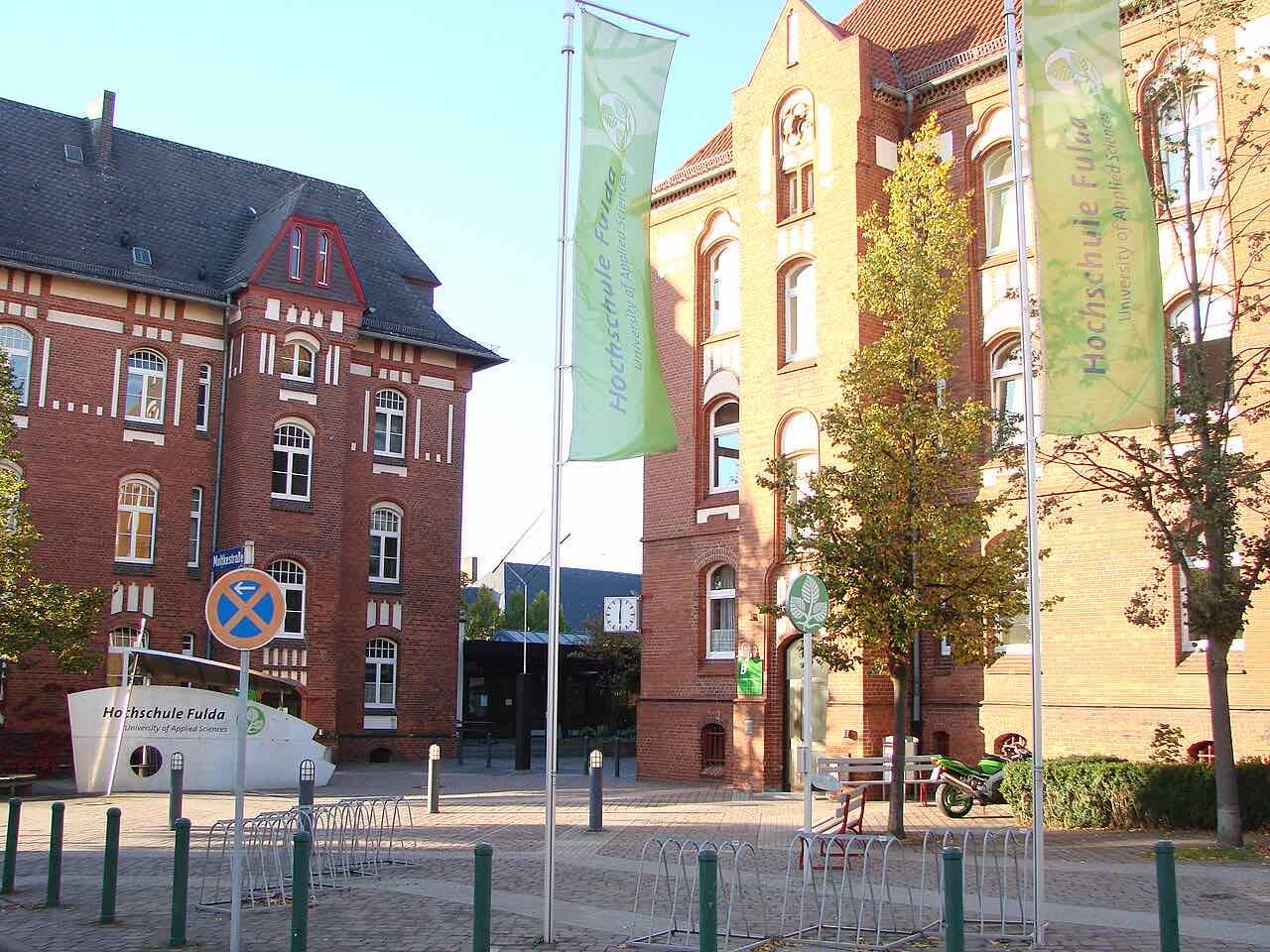 Red-brick campus buildings with green Hochschule Fulda University of Applied Sciences banners on flagpoles and bicycle racks in the foreground