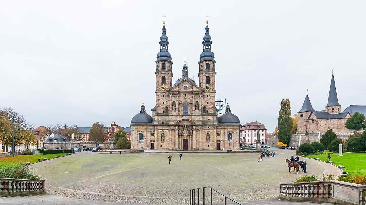 The twin-towered baroque cathedral of Fulda on its wide paved square, with the small stone Michaelskirche and a lawn on the right under a grey sky