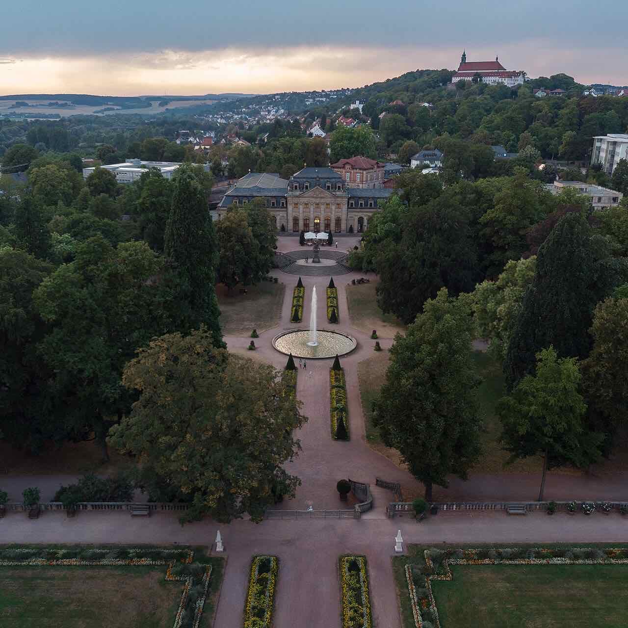 Aerial view at dusk of the Fulda palace garden with its straight avenues, a fountain and the Orangerie, and the Frauenberg monastery on the hill behind