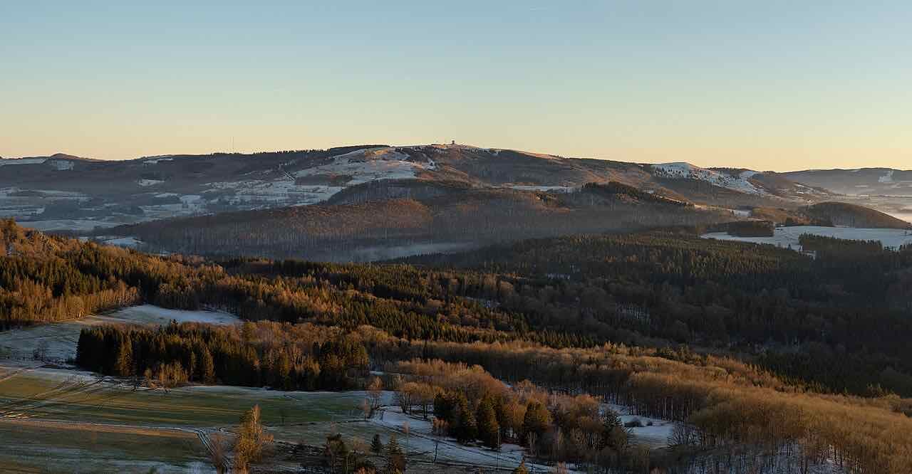 The rounded, open ridges of the Rhön in low winter light, with patches of snow on the high ground and dark forest in the valleys