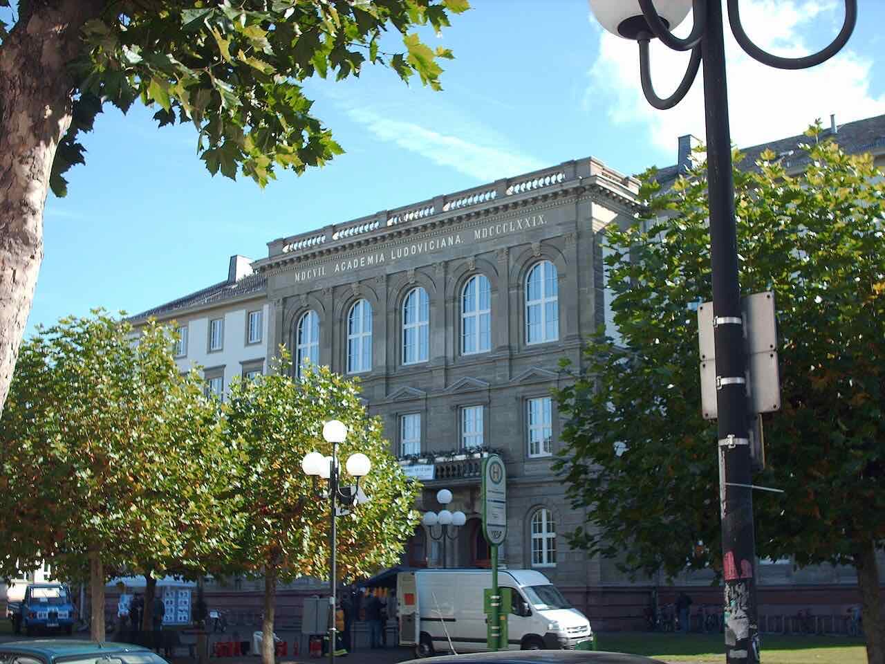 The sandstone main building of Justus Liebig University seen between plane trees, with the Latin inscription Academia Ludoviciana and the year 1607 along the top