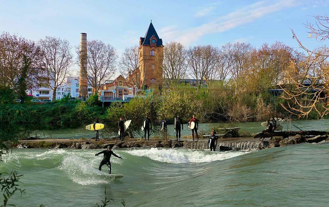 A surfer riding the standing wave on the Lahn while others in wetsuits wait on the weir, with a brick tower and chimney behind
