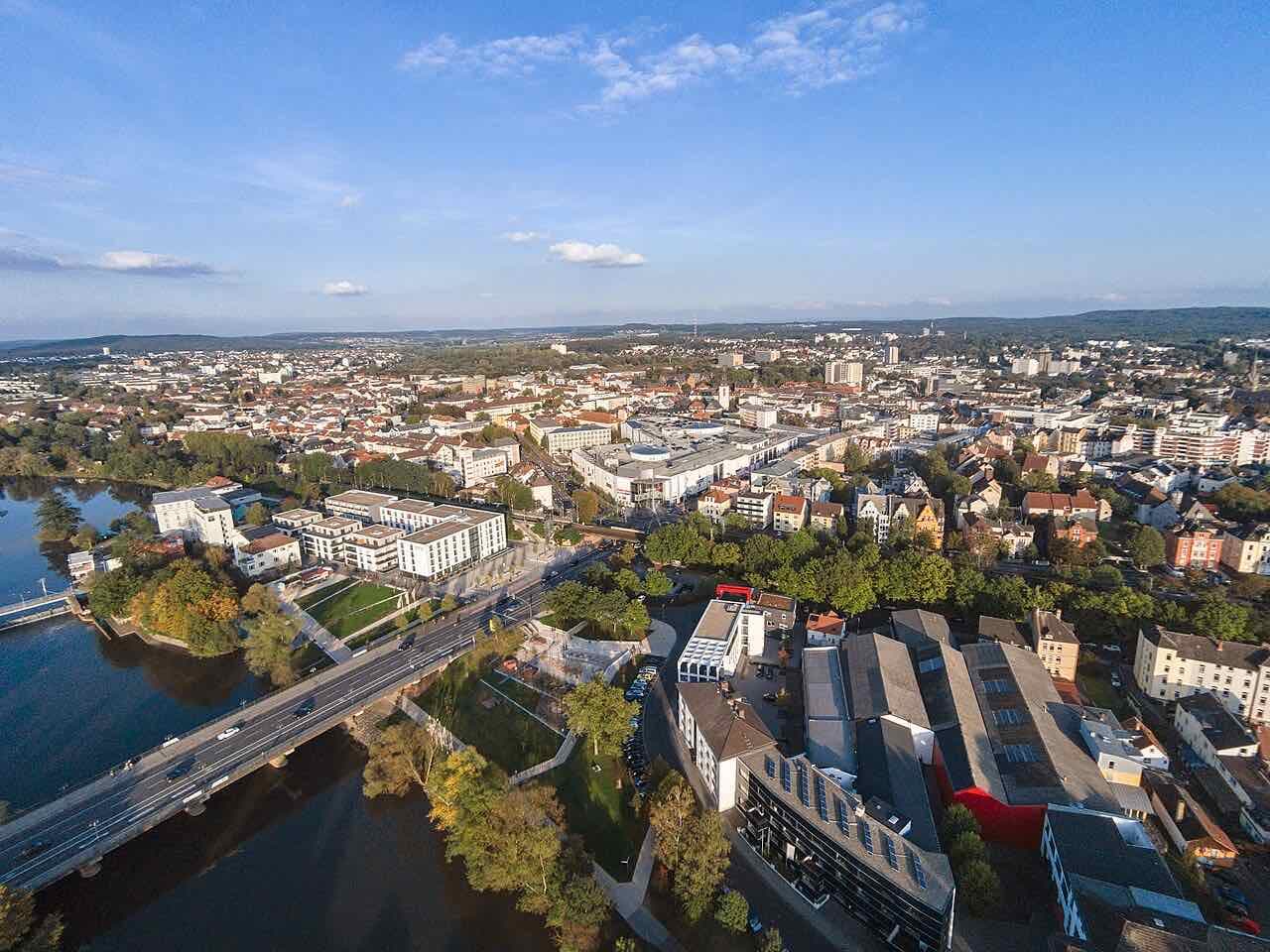 Aerial view of Gießen on a clear day, with the Lahn and a road bridge in the foreground, the shopping centre and the town's roofs behind, and wooded hills on the horizon