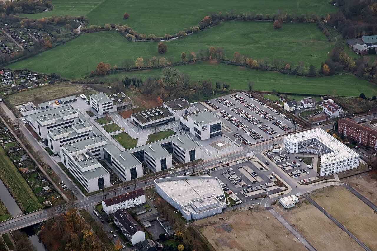 Aerial view of the Hamm campus of Hochschule Hamm-Lippstadt: pale modern buildings and car parks between fields and housing