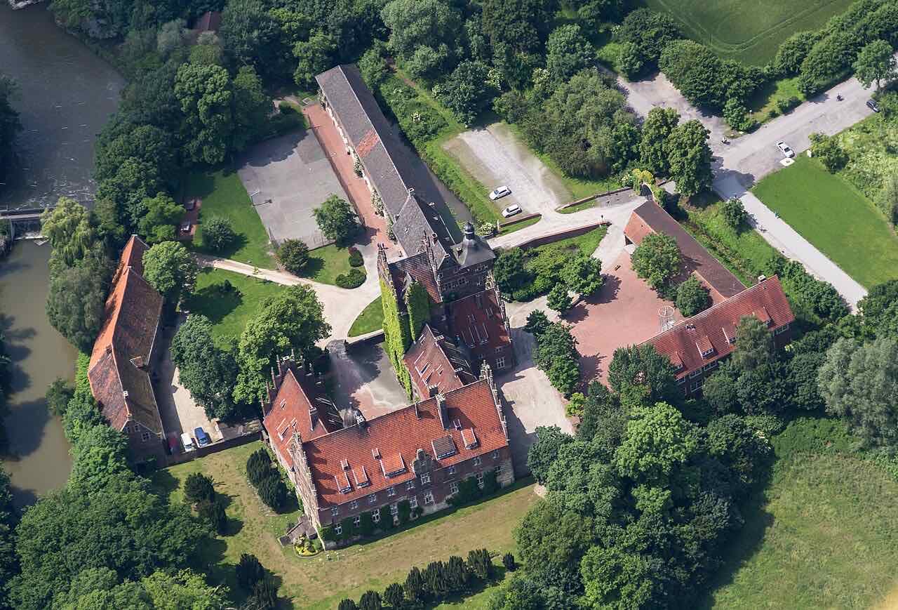 Aerial view of Schloss Heessen: red-roofed brick buildings around a courtyard, surrounded by trees and a river