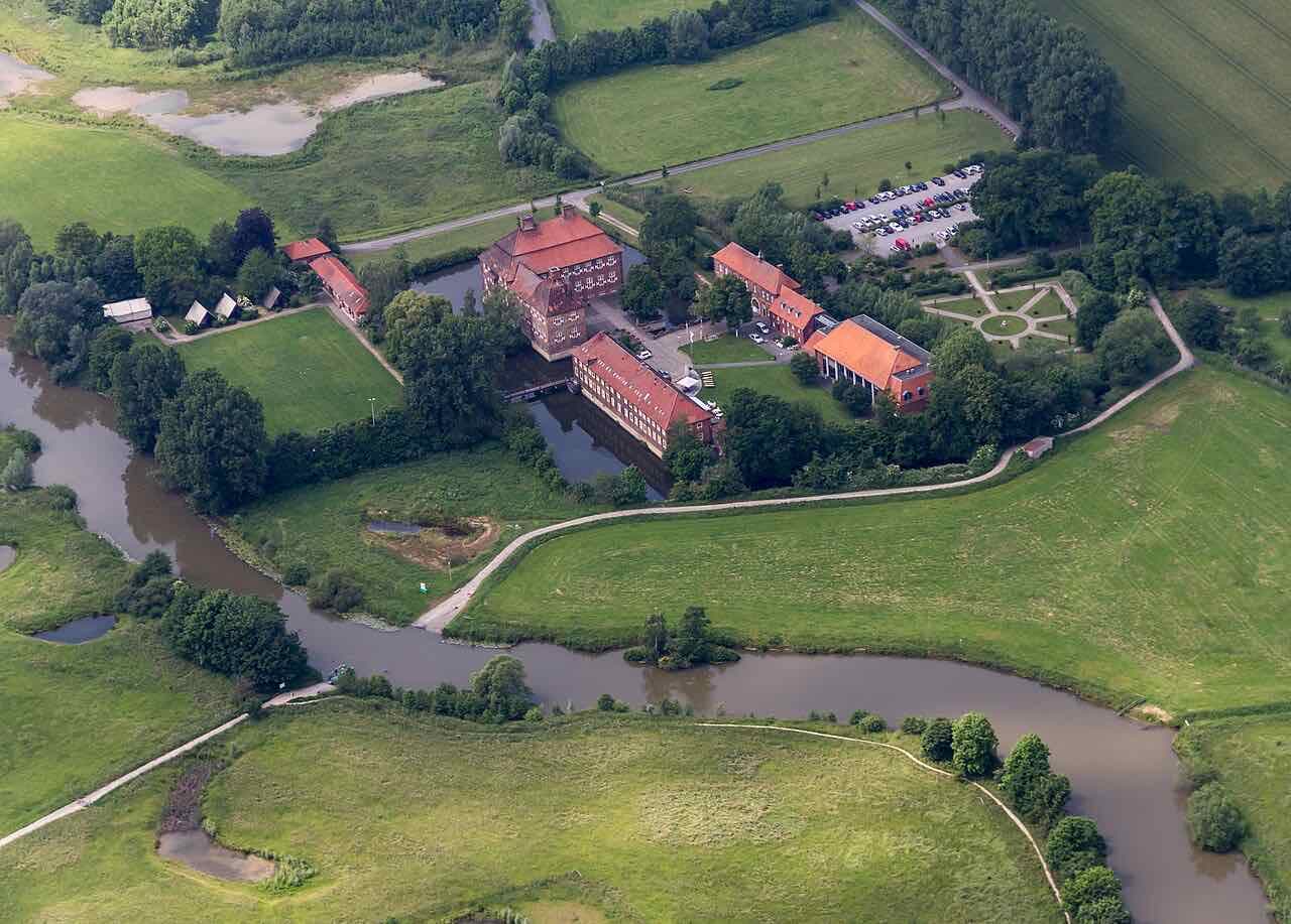 Aerial view of Schloss Oberwerries: red brick buildings with a water moat in green meadows beside a river bend