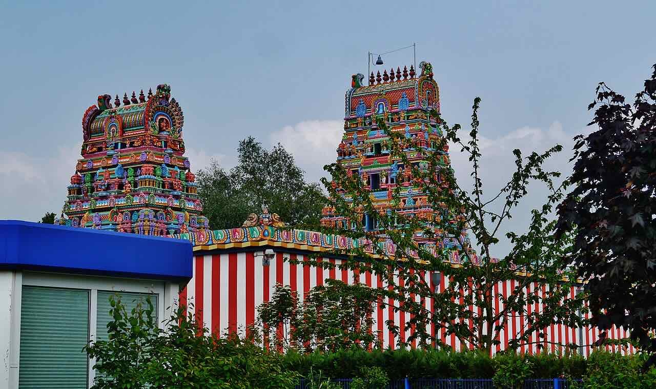 Two brightly painted temple towers rising above red and white striped walls and a hedge under a grey-blue sky