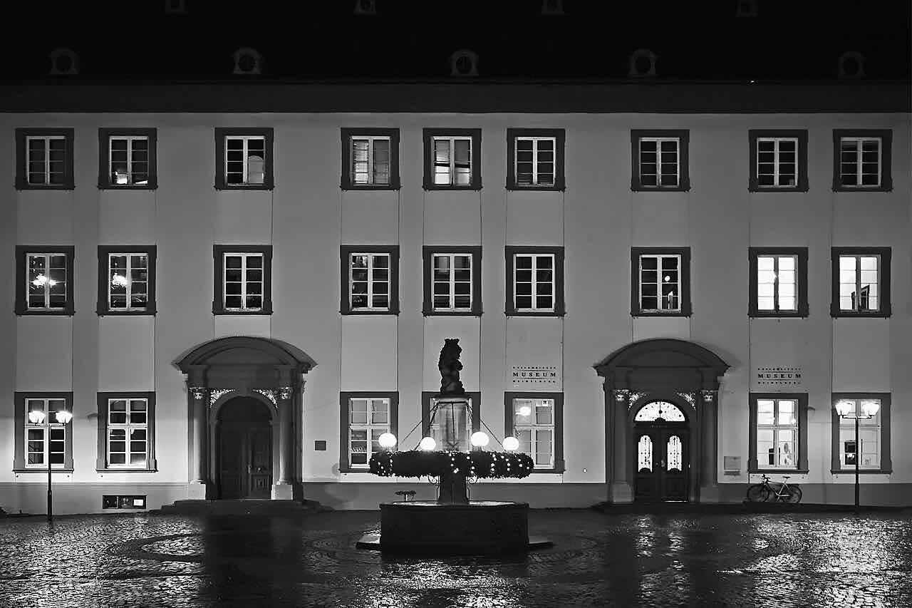 The pale facade of the Old University at night in black and white, with a lit fountain in the square in front