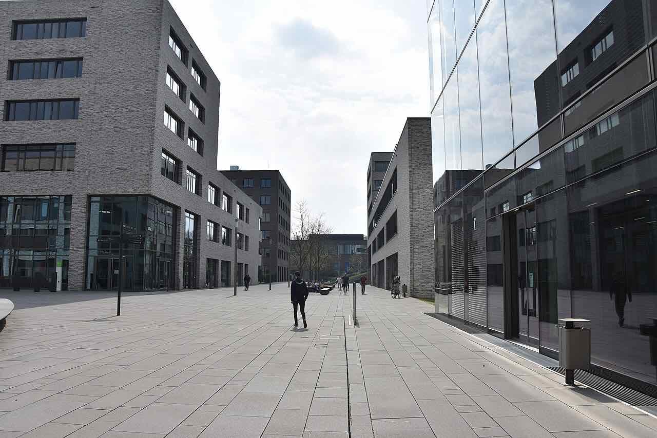 A paved walkway between brick-clad and glass university buildings on the Bildungscampus, with a few people walking and a bike parked