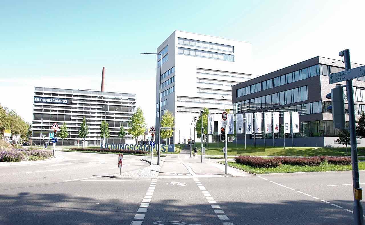 The Bildungscampus in Heilbronn: a white high-rise, a long grey building with the word Bildungscampus on its facade and a glass building with university banners, seen from a crossing on a sunny day